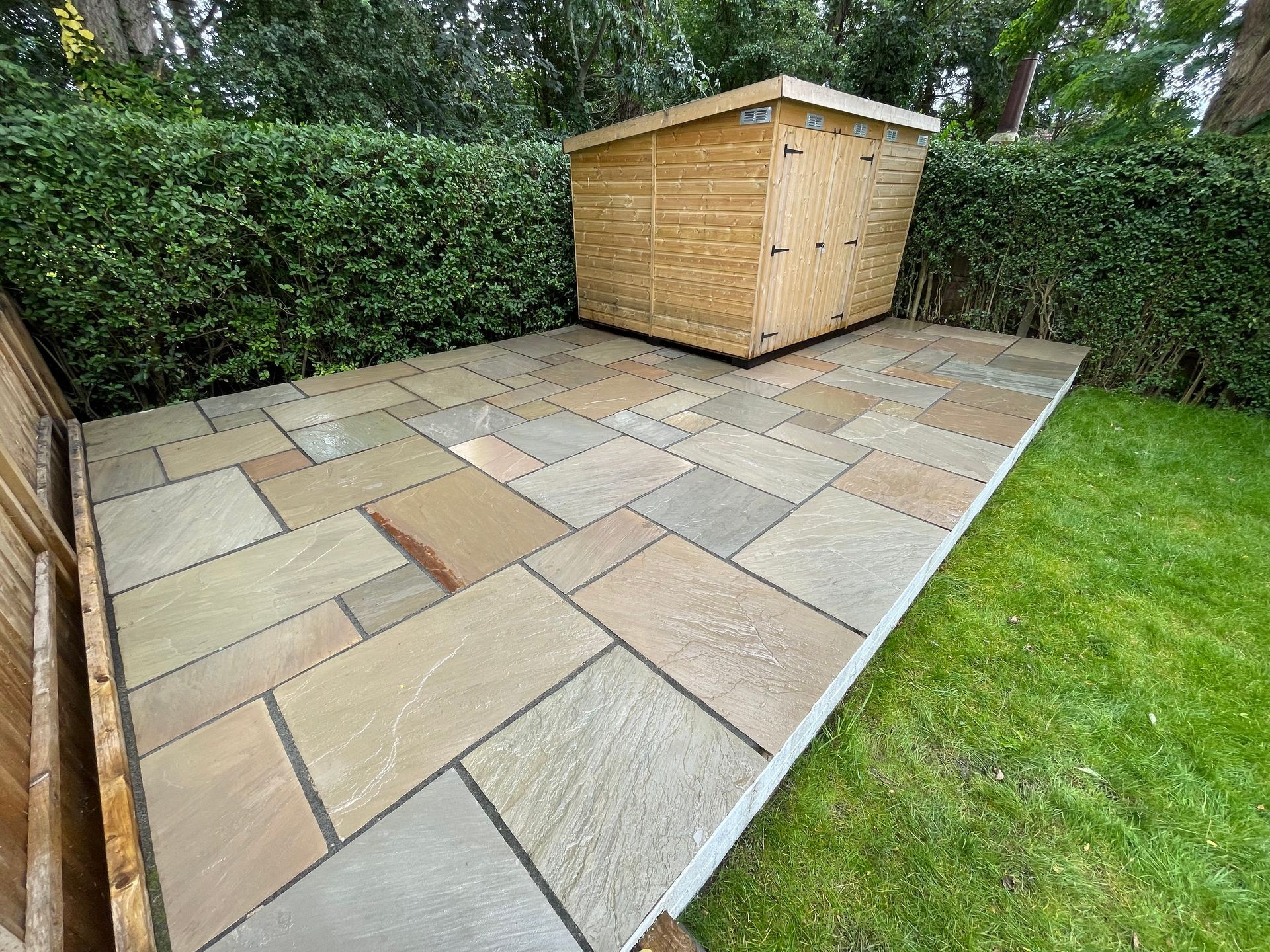 A wooden shed on a newly laid stone patio surrounded by green hedges and grass.