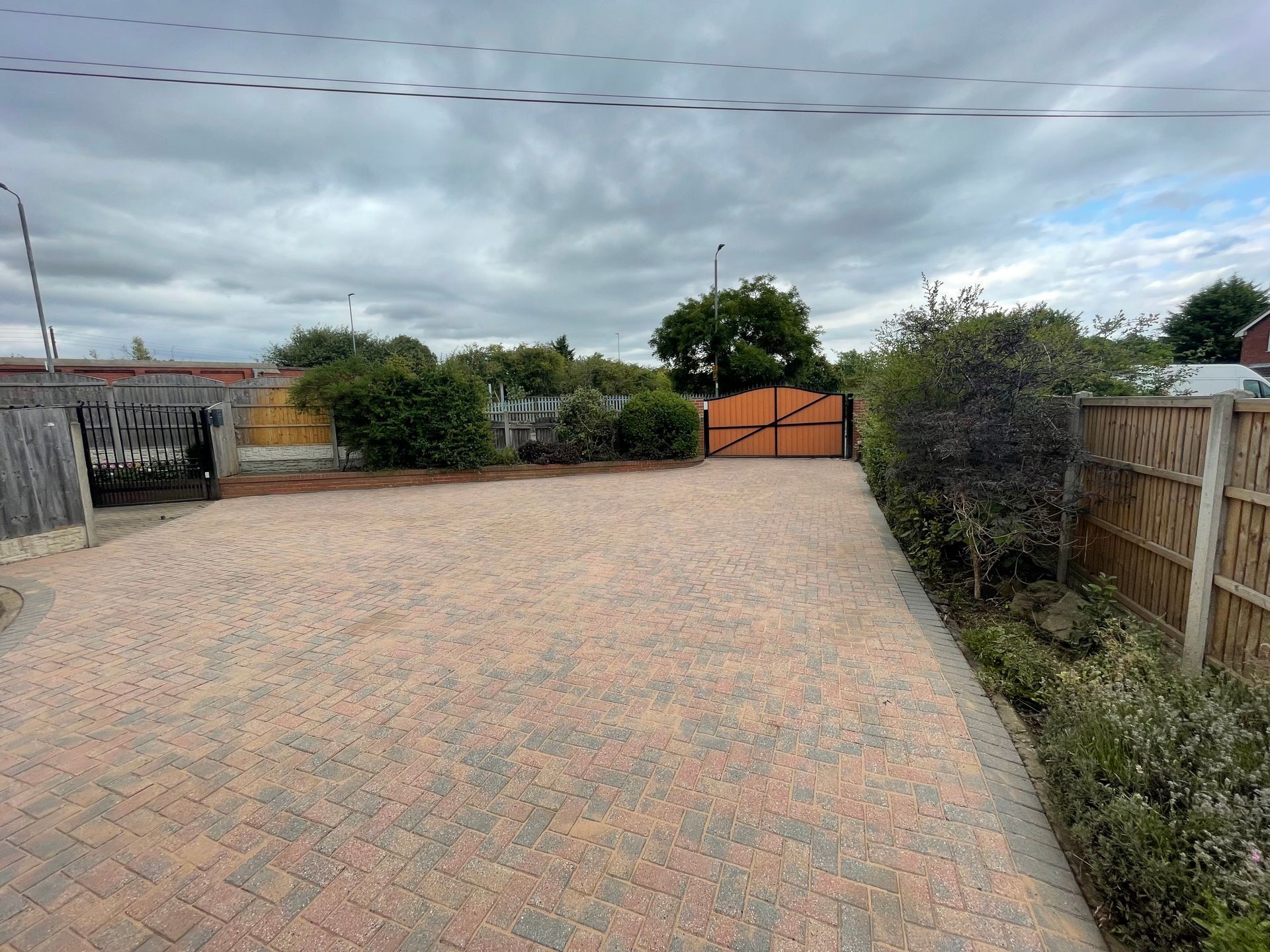 Spacious paved driveway with wooden gate, surrounded by wooden fences and greenery under a cloudy sky.